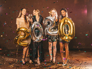 Fototapeta na wymiar Beautiful Women Celebrating New Year.Happy Gorgeous Girls In Stylish Sexy Party Dresses Holding Gold and Silver 2020 Balloons, Having Fun At New Year's Eve Party.Сarrying and drinking champagne flutes