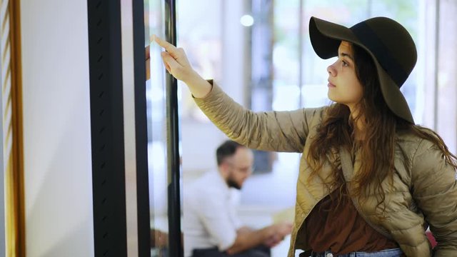 Female using touchscreen terminal in mall. Elegant young woman in hat consults on modern big timetable or info screen in interior. Side view girl touching display at out of focus background