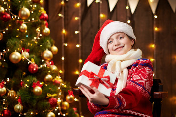 Cheerful santa helper girl with gift box sitting indoor near decorated xmas tree with lights, dressed in red sweater - Merry Christmas and Happy Holidays!