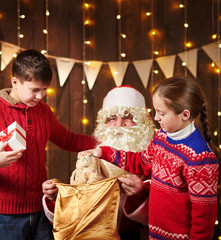 Santa Claus opens a bag and gives gifts to children, sitting indoor near decorated xmas tree with lights - Merry Christmas and Happy Holidays!