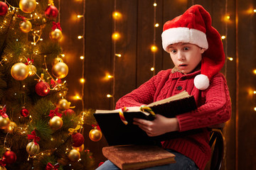 Teen boy reading book, sitting indoor near decorated xmas tree with lights, dressed as Santa helper - Merry Christmas and Happy Holidays!