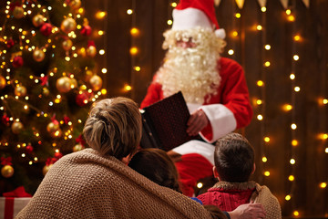 Santa Claus reading book for family. Mother and children sitting indoor near decorated xmas tree with lights - Merry Christmas and Happy Holidays!