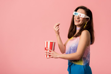 side view of happy young stylish disco girl in 3d glasses eating popcorn isolated on pink