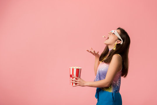 side view of happy young stylish disco girl in 3d glasses throwing popcorn in mouth isolated on pink