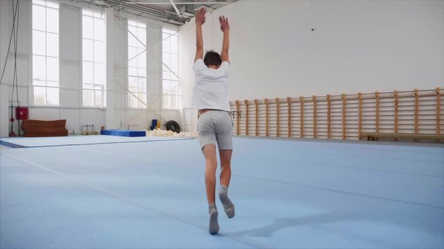 A teenager boy in a white T-shirt and grey shorts is doing a cartwheel and a triple back flip and lands on feet in gym on the blue sport mats, steadicam shot in slow motion.