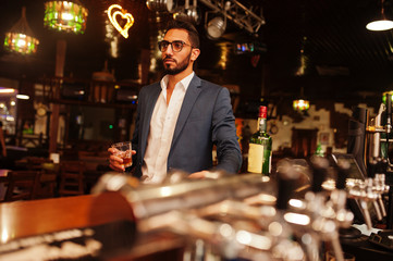 Handsome well-dressed arabian man with glass of whiskey and cigar posed at pub.