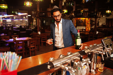 Handsome well-dressed arabian man with glass of whiskey and cigar posed at pub.