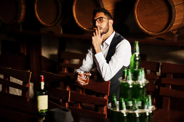 Handsome well-dressed arabian man with glass of whiskey and cigar posed at pub.