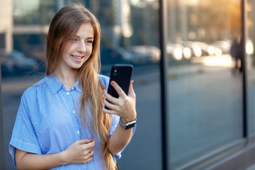 Happy smiling attractive young woman with long hair and smart watches using a smartphone near the windows of office building. Copy space. Businesswoman concept. Working female.