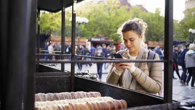 Female tourist walking at easter street market in european city. Girl taking photo on smartphone process of making traditional Czech bakery products trdelnik, prepared on old town square of Prague