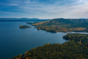Aerial drone view of Turgoyak lake in autumn surrounded by Ural mountains, hills, mixed forest with green pines and yellow birchs; colorful trees on coasts; fragile ecosystem and anthropogenic impact