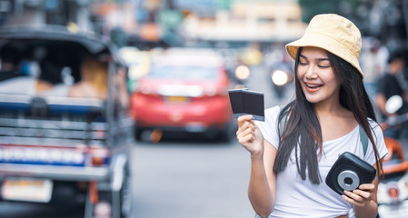 Fototapeta premium Traveller young Asian backpack woman wearing hat walking at Khao San road hands holding instant camera and film to take a picture, famous travelling landmark in Bangkok city of Thailand. 