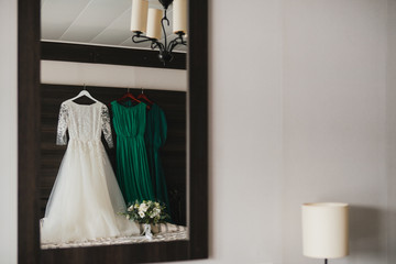 Modern bridal fashion. White wedding dress and green bridesmaids dresses hanging in the hotel room.