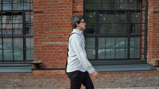 Middle gimbal profile shot. A serious young man walking fast from left to right along the street in eyeglasses wearing blue shirt on background of red brick building with windows