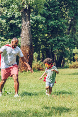  happy african american father and son running in costumes of superheroes in park