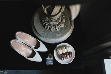 Botanic bridal chic. Female classic shoes, perfume bottle, macaron dessert, wedding rings and engagement ring on black background.