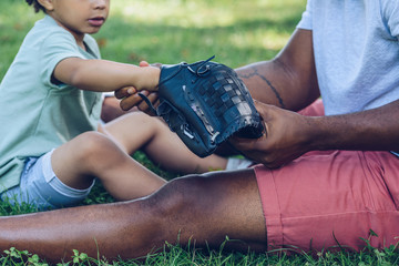 cropped view of african american man showing baseball glove to son while sitting on lawn in park