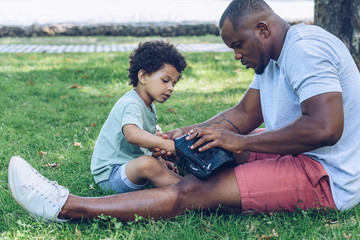 handsome african american man showing baseball glove to son while sitting on lawn in park