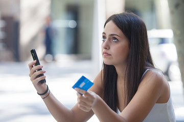 Worried young woman with credit card and phone sitting in a terrace bar