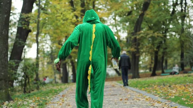 Back of green dinosaur male actorwalking slow in the park tottering carrying beer bottle. Drunken youth. Addictive generation.