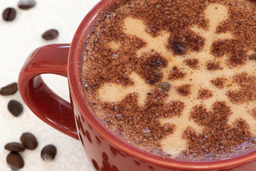 A foamy mug of cappuccino coffee with chocolate snowflake motif and a snowy background with a scattering of coffee beans sets the scene for a festive Christmas and warming up in cold winter weather