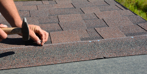 Roofer hands installing asphalt shingles on house construction roof corner with hammer and nails. ...