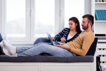 Young couple siting by the window and using at laptop spending time together at home.