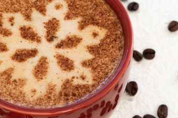A foamy mug of cappuccino coffee with chocolate snowflake motif and a snowy background with a scattering of coffee beans sets the scene for a festive Christmas and warming up in cold winter weather