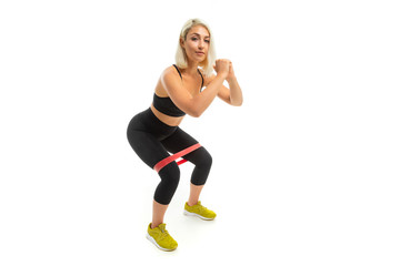 A young sports girl with blonde hair and bright manicure in black sports topics, leggings and sneakers does exercises with sports rubber.