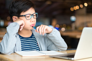 Smart looking Asian preteen boy holding pencil against his chin, thinking and looking at computer laptop. Researching, studying and solving problem with concentration. Online learning concept.