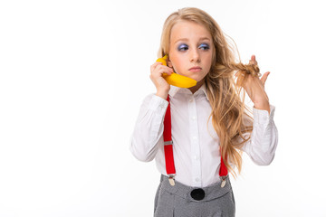 Little girl with long blonde hair in white shirt, red pull-ups, pants in cage, red socks and shoes with bright makeup with banana