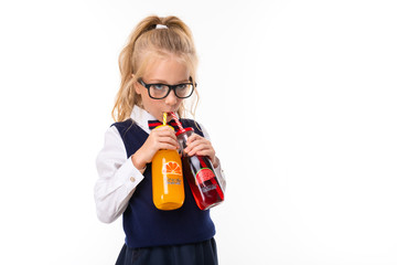 A little girl with blond hair stuffed in a horse tail, large blue eyes and a cute face in square black glasses drinks orange and watermelon juice in a glass bottle of tube.