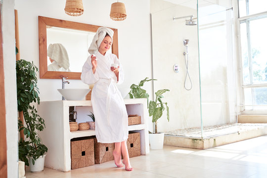Full-length photo of woman in bathrobe with cream in her hands in bathroom.