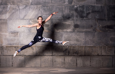 Fototapeta na wymiar Female dancer in black tank top and leggings jumping in front of a dark gray stone wall