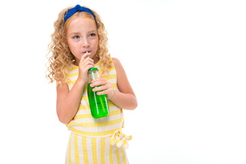 Obraz premium A little girl with red heap hair in a white and yellow striped summer suit, with a blue bandage on her head drinks orange juice in a glass bottle of pipe.