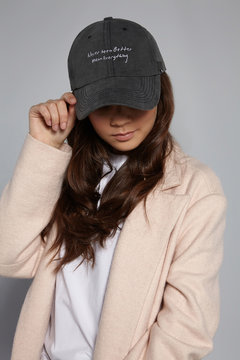 Medium close-up shot of brown-haired lady in a white top, beige jacket and a gray baseball cap with an embroidered lettering "Never been better mean everything" and decorated with a metal semi-ring.