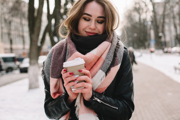 Happy young woman with disposable cup wearing scarf and leather jacket  while walking on city street during winter