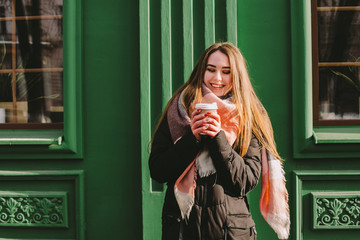Happy woman with disposable cup in warm clothing standing beside green building in city street during winter