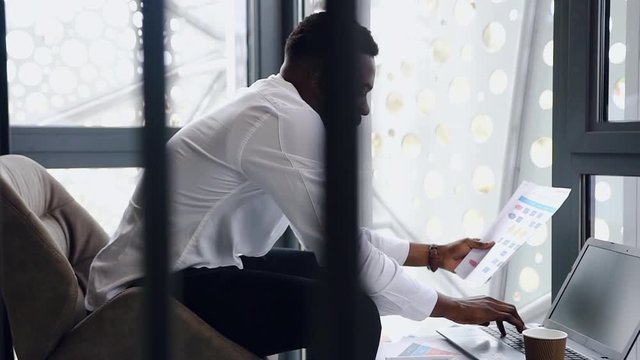 Close up of handsome careful african american manager entering datas from report with charts to the computer in specially designted room