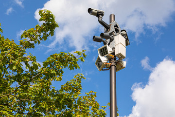 CCTV cameras and white street lamps on one column against sky and green leaves of maple tree