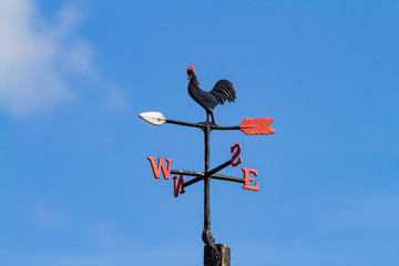 Cockerel Rooster weather vane, wind vane, weathercock against blue sky. Wind direction instrument, with letters for compass points, painted black red and white.