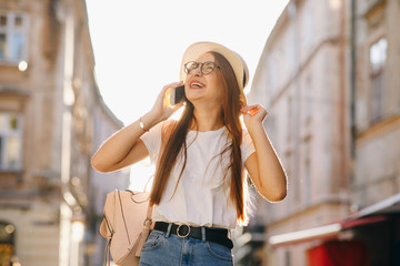 Attractive woman travels in hat and eyeglasses stands on city street, talking on cell phone, smiling, laughing. Hipster girl walks. Vacation, adventure, trip. Sunny day, backlight.