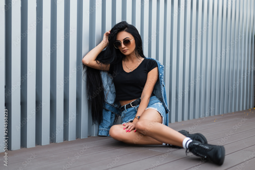 Wall mural Young hipster woman in sunglasses in a trendy blue denim jacket in stylish leather black autumn boots in a skirt is sitting near a silver metal wall on the street. American girl. Modern youth fashion.