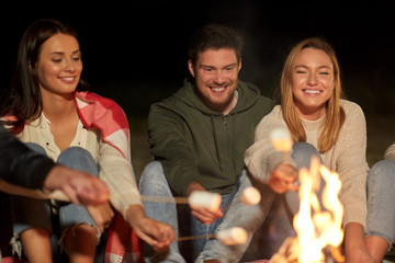 leisure and people concept - group of smiling friends sitting at camp fire and roasting marshmallow...