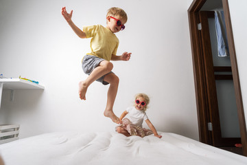 A little boy in red sunglasses and a yellow skirt jumps on a white mattress. Studio portrait on a white background.