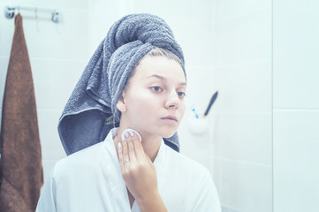 the girl in the bathroom in front of the mirror with a towel on her head applies cream to her face. selective focus. film grain