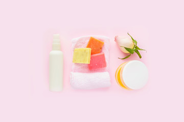 Open jar of cosmetic cream, white cosmetics bottle, soap, rose and towel on pink background. Concept of natural spa cosmetics. Flat lay, top view, copy space .