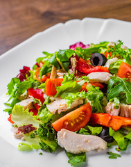 Fresh salad with chicken breast, arugula, black olives,red pepper, lettuce, fresh sald leaves and tomato on a white plate on wooden table background