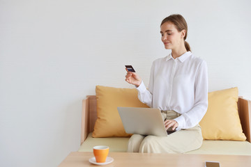 young attractive caucasian woman wearing white casual clothes shopping and payment online by using laptop computer, smart phone and credit card while drinking a cup coffee on table with relaxation. 