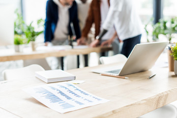 selective focus of desk with laptop, notebook and paper with graphs and charts near businesspeople standing at workplace in office